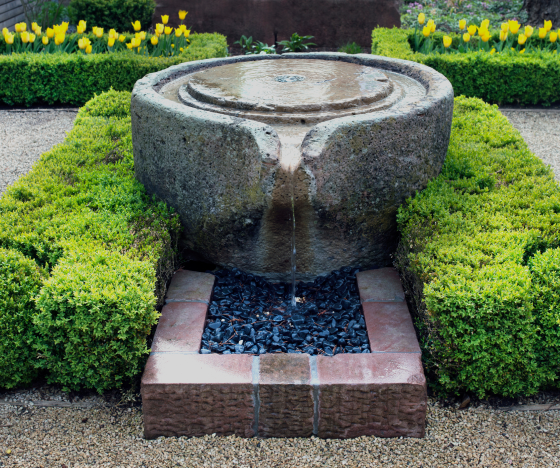 a stone fountain surrounded by green bushes and flowers.